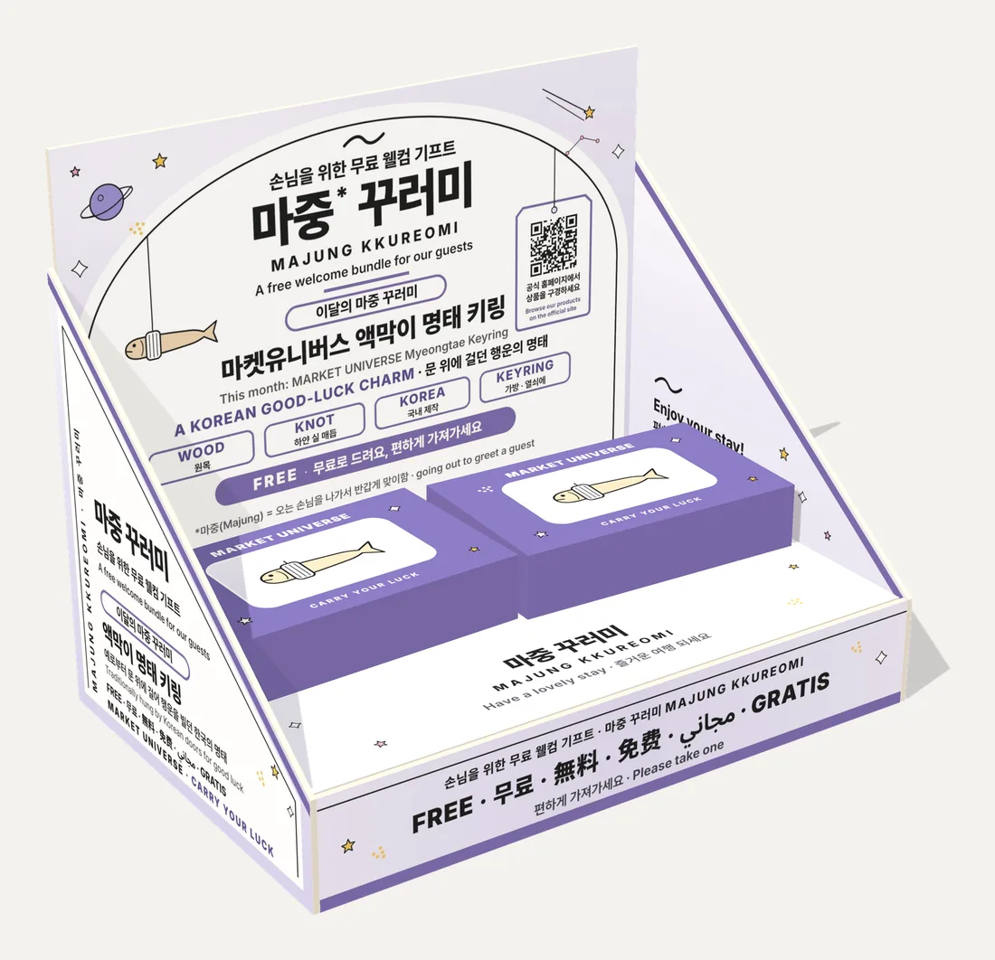 마켓유니버스 액막이 명태 키링 마중 꾸러미 매대 3D 시안. 라벤더색 매대 정면에 명태 그림과 QR이 있고 보라색 상자 두 개가 담겨 있다.